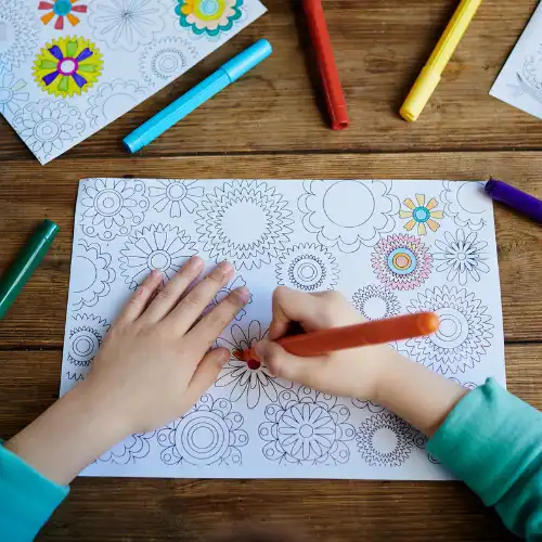 Close up of a child's hands coloring a complex pattern worksheet with markers.